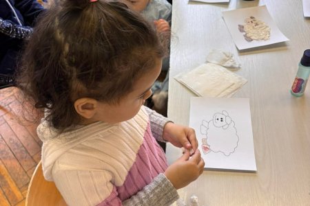 A képen a gyerekek egy asztalnál az előre megrajzolt bárányra ragasztják a papírzsebkendőből gyúrt galacsinokat.